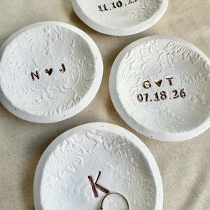 Puede incluir: Cuatro cuencos de cer&aacute;mica blanca para anillos con patrones florales en relieve. Tres cuencos tienen iniciales y fechas en letras color cobre. Un cuenco contiene un anillo de oro. Las fechas son 11.10.25, 07.18.26.