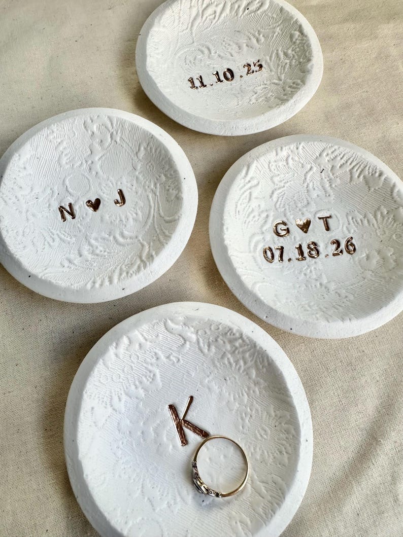 Puede incluir: Cuatro cuencos de cer&aacute;mica blanca para anillos con patrones florales en relieve. Tres cuencos tienen iniciales y fechas en letras color cobre. Un cuenco contiene un anillo de oro. Las fechas son 11.10.25, 07.18.26.