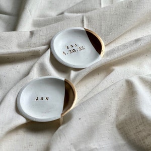 May include: Two white ceramic ring dishes with gold accents. One dish has the inscription "E&J 4.20.22" and the other has the inscription "J&N".