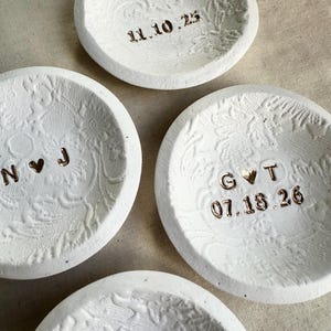 Puede incluir: Cuatro peque&ntilde;os cuencos de cer&aacute;mica blanca con patrones florales en relieve. Cada cuenco tiene una fecha o iniciales diferentes en color dorado met&aacute;lico. Las fechas son 11.10.25 y 07.18.26. Las iniciales son N♡J y G♡T.