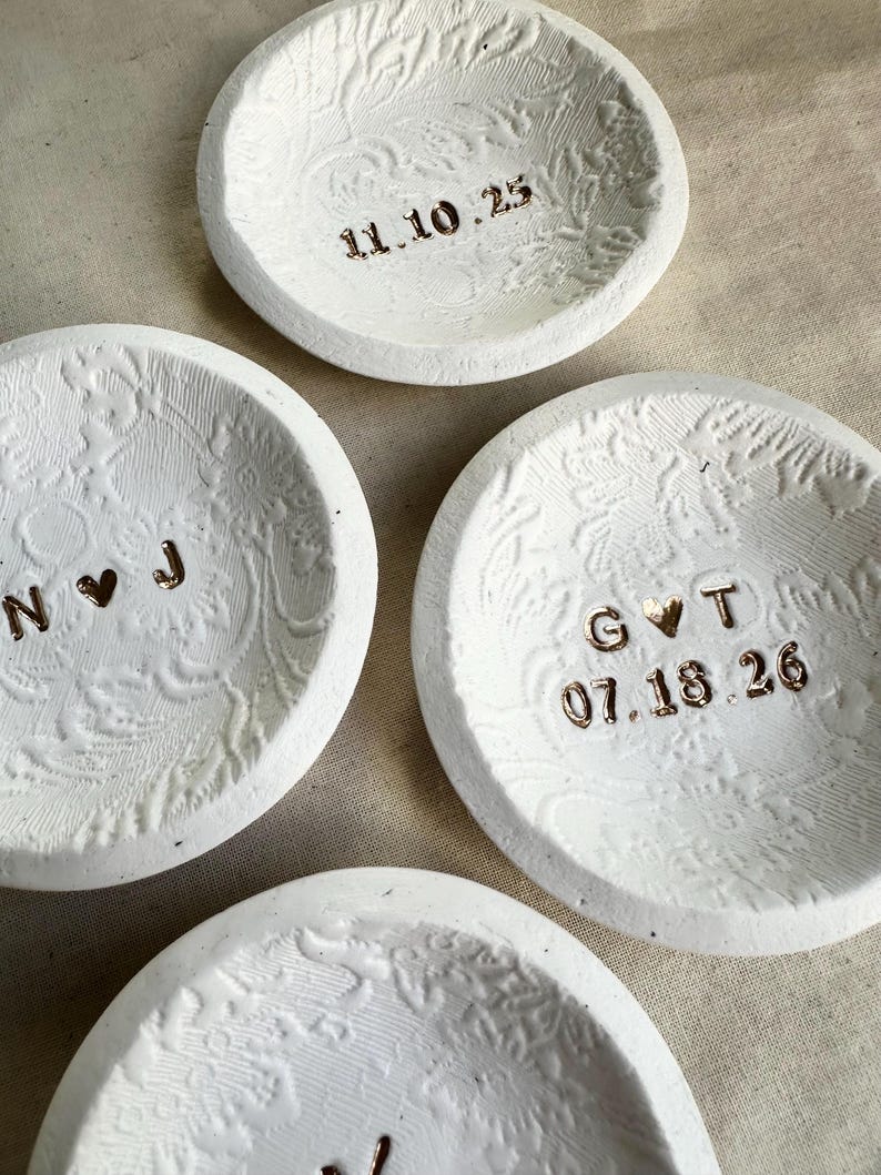 Puede incluir: Cuatro peque&ntilde;os cuencos de cer&aacute;mica blanca con patrones florales en relieve. Cada cuenco tiene una fecha o iniciales diferentes en color dorado met&aacute;lico. Las fechas son 11.10.25 y 07.18.26. Las iniciales son N♡J y G♡T.