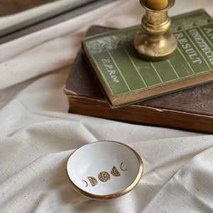 K&ouml;nnte beinhalten: Eine wei&szlig;e Keramik-Ring Schale mit goldfarbenem Rand und einem Design aus f&uuml;nf goldenen Glitzermonden. Die Schale steht auf einer wei&szlig;en Stoffoberfl&auml;che.