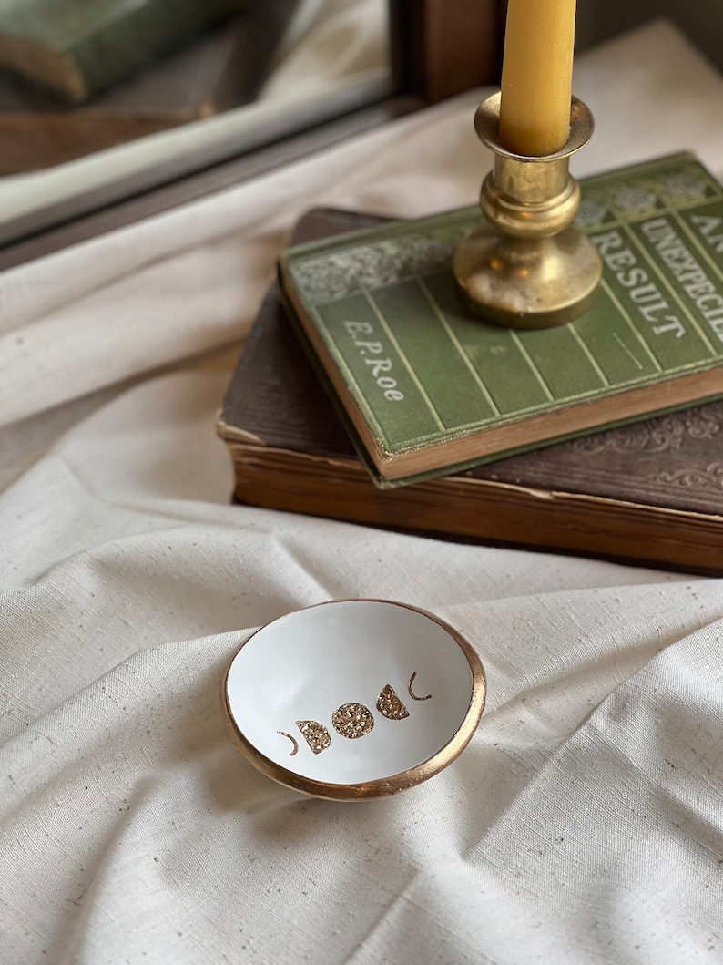 K&ouml;nnte beinhalten: Eine wei&szlig;e Keramik-Ring Schale mit goldfarbenem Rand und einem Design aus f&uuml;nf goldenen Glitzermonden. Die Schale steht auf einer wei&szlig;en Stoffoberfl&auml;che.