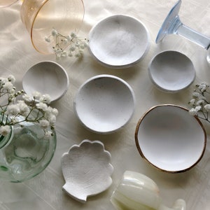 May include: A set of white ceramic dishes with a variety of shapes and sizes. The dishes are arranged on a white surface with a few sprigs of white flowers.