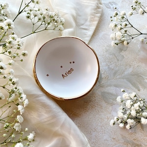 Puede incluir: Plato de cer&aacute;mica blanco para anillos con borde dorado y la palabra "Aries" escrita en letras doradas. El plato est&aacute; sobre un fondo de tela blanca con flores blancas.