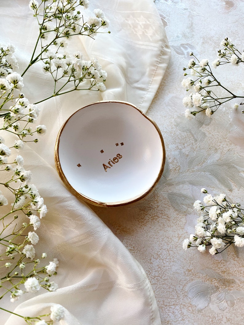 Puede incluir: Plato de cer&aacute;mica blanco para anillos con borde dorado y la palabra "Aries" escrita en letras doradas. El plato est&aacute; sobre un fondo de tela blanca con flores blancas.