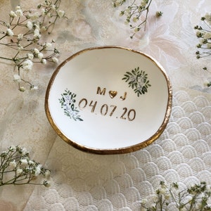Puede incluir: Plato de cer&aacute;mica blanca para anillos con borde dorado y dise&ntilde;o pintado en oro de hojas y flores. El plato tiene las iniciales "M & J" y la fecha "04.07.20" escritas en oro.