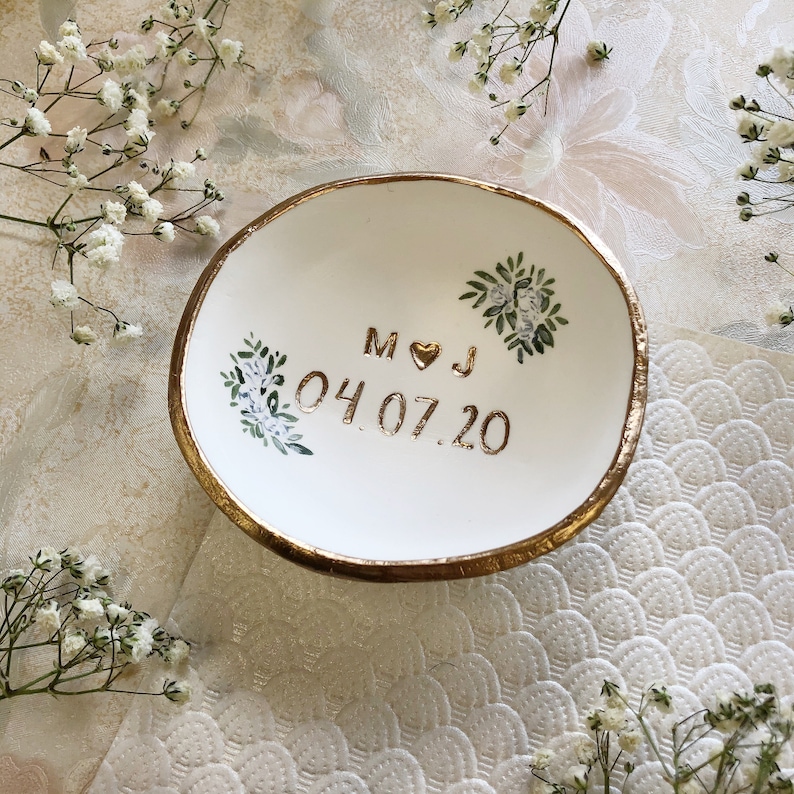 Puede incluir: Plato de cer&aacute;mica blanca para anillos con borde dorado y dise&ntilde;o pintado en oro de hojas y flores. El plato tiene las iniciales "M & J" y la fecha "04.07.20" escritas en oro.