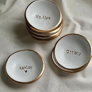 May include: Three white ceramic ring dishes with gold trim. The dishes are personalised with the names "Ms. Alex", "Ashley", and "Christy".