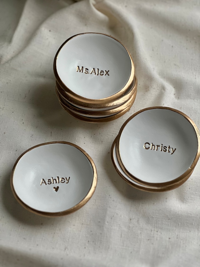 May include: Three white ceramic ring dishes with gold trim. The dishes are personalised with the names "Ms. Alex", "Ashley", and "Christy".