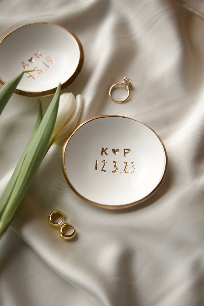 May include: Two white ceramic ring dishes with gold trim. The top dish has the text "A&K 12.8.19" and the bottom dish has the text "K&hearts;P 12.3.23".