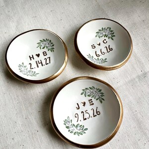 May include: Three white ceramic ring dishes with gold rims. Each dish features a floral design and personalised text with initials and dates in brown. The dishes are arranged on a neutral-coloured surface.