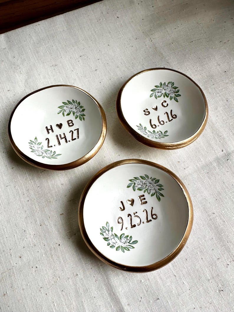 May include: Three white ceramic ring dishes with gold rims. Each dish features a floral design and personalised text with initials and dates in brown. The dishes are arranged on a neutral-coloured surface.