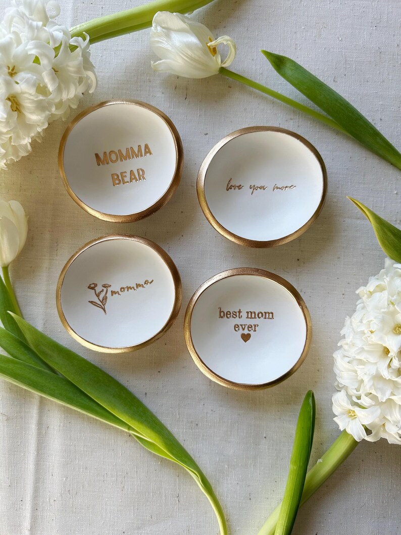 4 hand stamped white ring dishes, made by The Painted Press. One says "MOMMA BEAR", one says "momma" with a flower, one says "best mom ever" with a heart, and one says "love you more" in cursive. The words and rims of each dish are painted in gold.