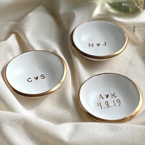 May include: Three white ceramic ring dishes with gold rims. Each dish has a different set of initials and a date stamped in the centre. The dishes are on cream-coloured fabric.