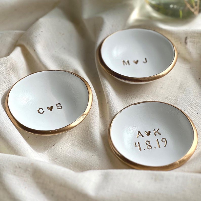 May include: Three white ceramic ring dishes with gold rims. Each dish has a different set of initials and a date stamped in the centre. The dishes are on cream-coloured fabric.