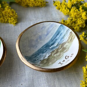 May include: A small, round, hand-painted ceramic dish with a gold rim. The interior features a watercolor painting of a beach scene with blue water and a sandy shore. The initials "I♡G" and the date "03.25" are painted on the dish.