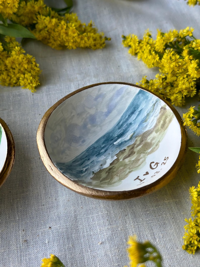 May include: A small, round, hand-painted ceramic dish with a gold rim. The interior features a watercolor painting of a beach scene with blue water and a sandy shore. The initials "I♡G" and the date "03.25" are painted on the dish.