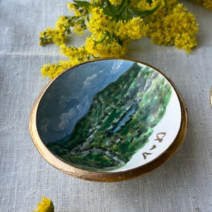 May include: A small, round, hand-painted ceramic bowl with a gold rim. The interior features a landscape painting of a mountain and sky. The initials "A <3 D" are painted on the white rim. Yellow flowers are in the background.