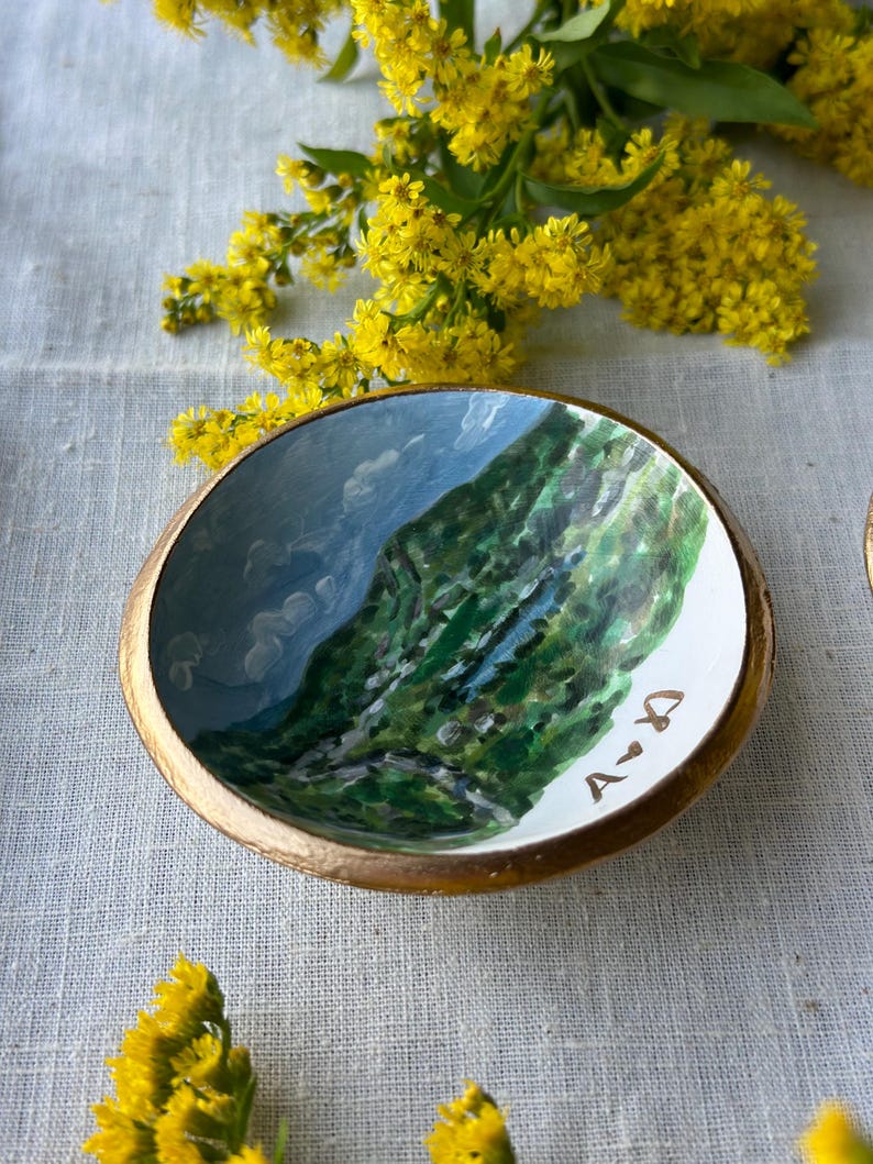 May include: A small, round, hand-painted ceramic bowl with a gold rim. The interior features a landscape painting of a mountain and sky. The initials "A <3 D" are painted on the white rim. Yellow flowers are in the background.
