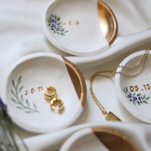 May include: Four white ceramic ring dishes with gold accents. Each dish has a different design, including initials, a date, and floral patterns. The dishes are on a white fabric background.