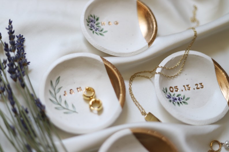 May include: Four white ceramic ring dishes with gold accents. Each dish has a different design, including initials, a date, and floral patterns. The dishes are on a white fabric background.