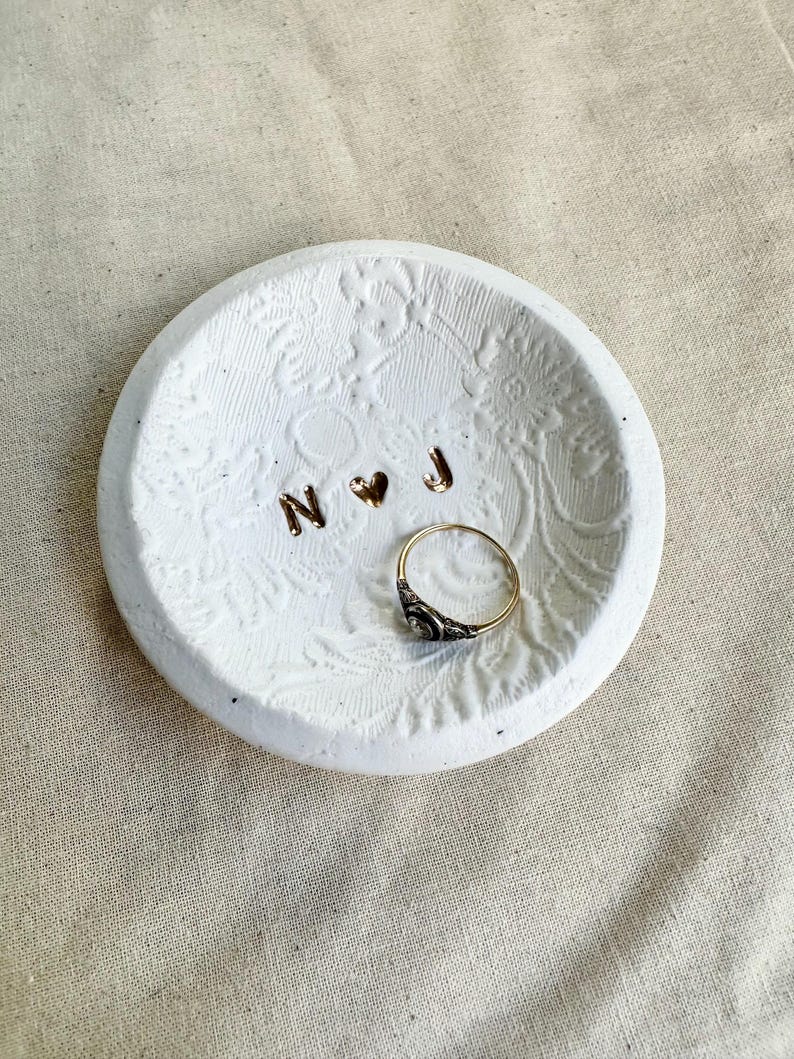 Puede incluir: Un plato redondo de cer&aacute;mica blanca con patrones florales en relieve. El plato contiene un anillo dorado y letras doradas que deletrean "N" y "J" con un s&iacute;mbolo de coraz&oacute;n. El anillo tiene un dise&ntilde;o negro y dorado.