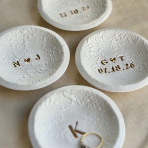 Puede incluir: Cuatro cuencos de cer&aacute;mica blanca para anillos con patrones florales en relieve. Uno de los cuencos contiene un anillo de oro. Cada cuenco tiene letras doradas: iniciales, una fecha o la letra "K". Los cuencos son peque&ntilde;os y redondos.