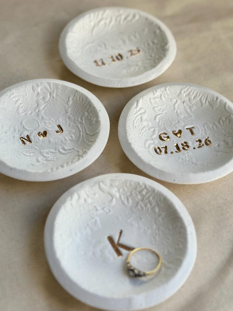 Puede incluir: Cuatro cuencos de cer&aacute;mica blanca para anillos con patrones florales en relieve. Uno de los cuencos contiene un anillo de oro. Cada cuenco tiene letras doradas: iniciales, una fecha o la letra "K". Los cuencos son peque&ntilde;os y redondos.