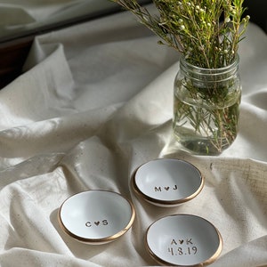 May include: Three white ceramic ring dishes with gold rims. The dishes are personalised with initials and a heart symbol. One dish has the initials "C & S", another has "M & J", and the third has "A & K" with the date "4.8.19".