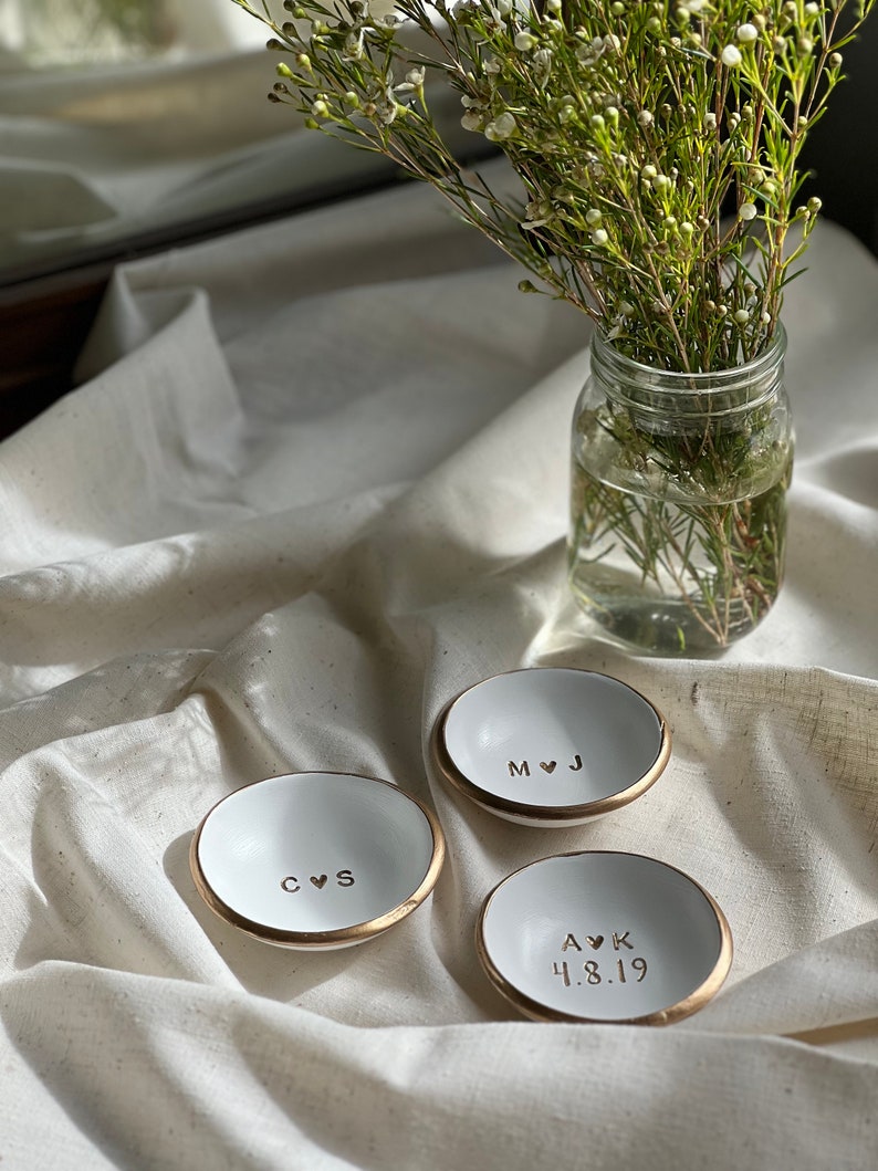 May include: Three white ceramic ring dishes with gold rims. The dishes are personalised with initials and a heart symbol. One dish has the initials "C & S", another has "M & J", and the third has "A & K" with the date "4.8.19".