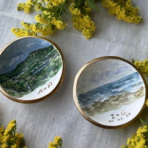 May include: Two small, round, hand-painted ceramic dishes with gold rims. One depicts a green and blue landscape, the other a beach scene. The first dish has the initials "A & D", the second "I & G 07.03.25".