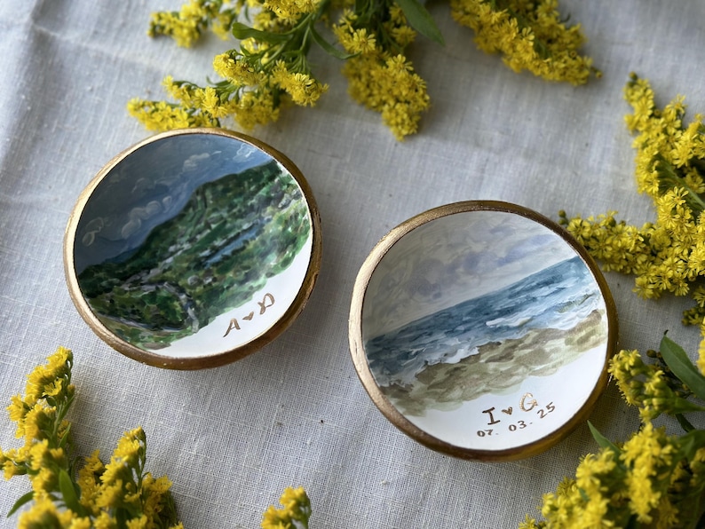 May include: Two small, round, hand-painted ceramic dishes with gold rims. One depicts a green and blue landscape, the other a beach scene. The first dish has the initials "A & D", the second "I & G 07.03.25".