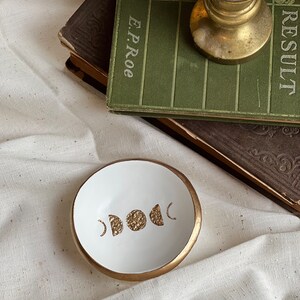 K&ouml;nnte beinhalten: Wei&szlig;e Keramik-Ring Schale mit goldfarbenem Rand und goldfarbenem Design aus Halbmonden und Vollmond.