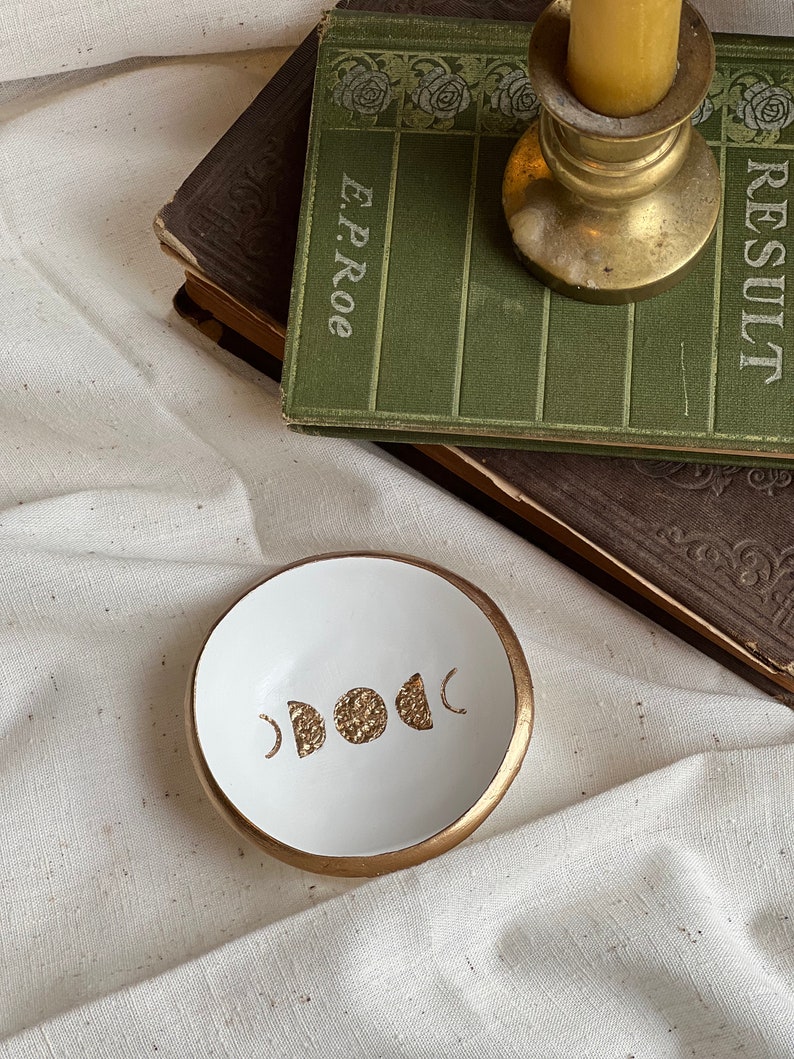 K&ouml;nnte beinhalten: Wei&szlig;e Keramik-Ring Schale mit goldfarbenem Rand und goldfarbenem Design aus Halbmonden und Vollmond.