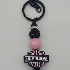 May include: Black and pink Harley-Davidson keychain. The keychain features a black clasp and ring, with a beaded design of black and pink beads. The Harley-Davidson logo is in pink and black.