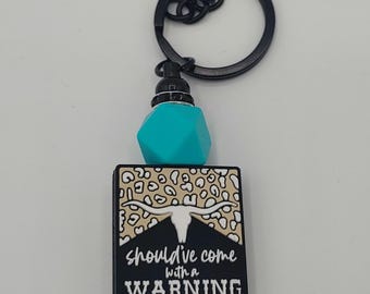 Può includere: Un portachiavi nero con un ciondolo rettangolare che recita "should've come with a WARNING". Il ciondolo ha un design a teschio di mucca e uno sfondo con stampa leopardata. Presenta anche un cubo turchese e un fondo nero perlato.