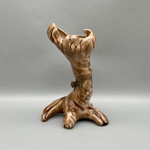 May include: A ceramic vase shaped like a tree trunk with a brown and white glaze. The vase has a small pine cone detail on the side.