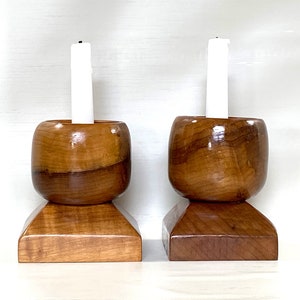 May include: A pair of wooden candle holders with a rounded top and square base. Each holder has a white candle.