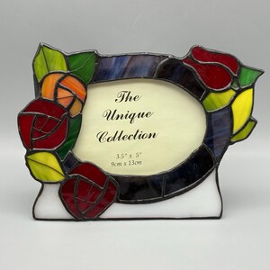 May include: A stained glass photo frame with a floral rose design. The frame is oval-shaped and features red, orange, yellow, and green roses. The text "The Unique Collection" is visible. The frame measures 3.5" x 5".