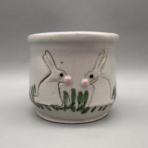 May include: A white ceramic planter with two outlined bunny figures and green grass detailing. The bunnies have pink noses and black eyes. The planter is cylindrical and has a smooth, unglazed finish.