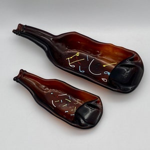 May include: Two brown glass bottle-shaped dishes. The larger dish has white, yellow, and blue accents. The smaller dish has white, yellow, and red accents. The dishes are made from recycled glass and are suitable for serving snacks or as decorative pieces.