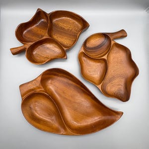 May include: Three wooden leaf-shaped serving trays. The trays are made of a warm-toned wood and feature multiple compartments. The largest tray has a long, curved leaf shape, while the others have more rounded forms. The trays are ideal for serving snacks or appetizers.