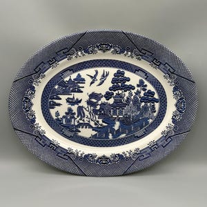 May include: An oval ceramic serving platter featuring a blue and white willow pattern. The central design depicts a landscape with a pagoda, trees, a bridge, and two birds in flight. The wide border has a geometric lattice and floral motif.