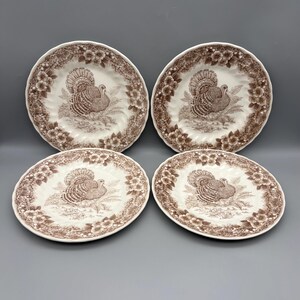 May include: A set of four round ceramic plates featuring a brown and white transferware pattern. Each plate displays a central illustration of a turkey surrounded by a detailed border of flowers and foliage with a scalloped edge design.