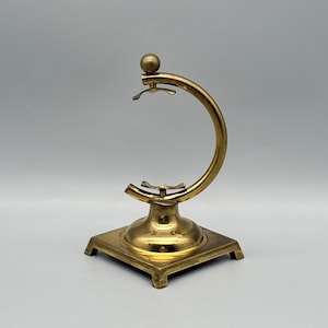 May include: A brass-colored, decorative stand with a square base and a curved arm. The arm has a small ball at the top and a hook-like feature. The stand is designed to hold an item.