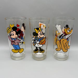 May include: Three clear drinking glasses featuring Disney characters Mickey Mouse, Minnie Mouse, and Pluto. Mickey is holding flowers, Minnie is holding a pie, and Pluto is holding a scroll. Each glass has the character's name printed on it.