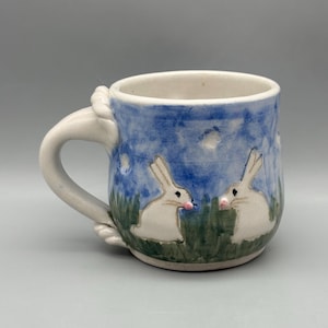 May include: Ceramic mug with a white handle and a blue and white painted exterior. The mug features two white rabbits with pink noses in a grassy field under a blue sky. The handle has a rope-like design.