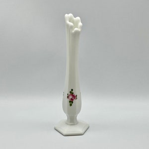 May include: A tall, white ceramic vase with a fluted rim and a hexagonal base. The vase features a delicate floral design of pink roses and green leaves. The vase is approximately 10 inches tall.