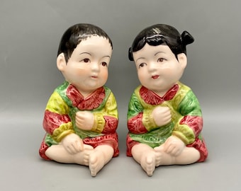 May include: A pair of ceramic figurines of children in traditional clothing. The boy is wearing a green, yellow, and red outfit, and the girl is wearing a green, red, and yellow outfit. Both figurines are sitting with their hands clasped in their laps.